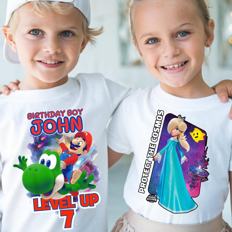May include: Two children wearing white t-shirts with colorful graphic designs. One shirt says "BIRTHDAY BOY JOHN" with Mario and Yoshi. The other shirt says "PROTECT THE COSMOS" with a princess character. Both shirts have a "Mario Galaxy" logo.