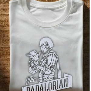 May include: White t-shirt with a grey and white graphic of The Mandalorian and Grogu. The text "The Mandalorian" is at the bottom. The shirt is folded, showing the crew neck and the printed design on the front.