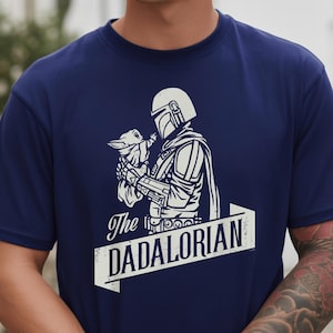 May include: Navy blue t-shirt with a white graphic of a character in armour holding a small creature. The text "The DADALORIAN" is displayed in a white banner below the image. A unique design for fans of the series.