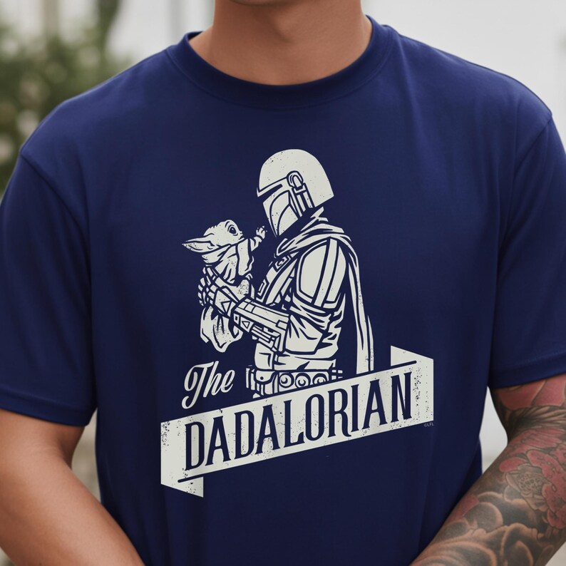 May include: Navy blue t-shirt with a white graphic of a character in armour holding a small creature. The text "The DADALORIAN" is displayed in a white banner below the image. A unique design for fans of the series.