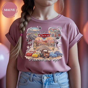 Puede incluir: Camiseta de color malva con un gr&aacute;fico colorido de los personajes de la pel&iacute;cula Cars. El dise&ntilde;o incluye el texto "2026 Cars Family Trip" e im&aacute;genes de circuitos y una gasolinera. La camiseta tiene las mangas dobladas.