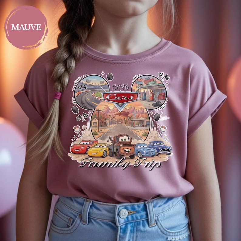 Puede incluir: Camiseta de color malva con un gr&aacute;fico colorido de los personajes de la pel&iacute;cula Cars. El dise&ntilde;o incluye el texto "2026 Cars Family Trip" e im&aacute;genes de circuitos y una gasolinera. La camiseta tiene las mangas dobladas.