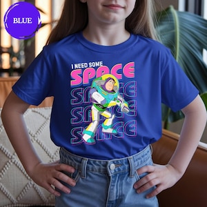 May include: Blue t-shirt featuring a graphic of Buzz Lightyear with the text "I NEED SOME SPACE". The design includes the word "SPACE" repeated vertically. The shirt is worn by a person.