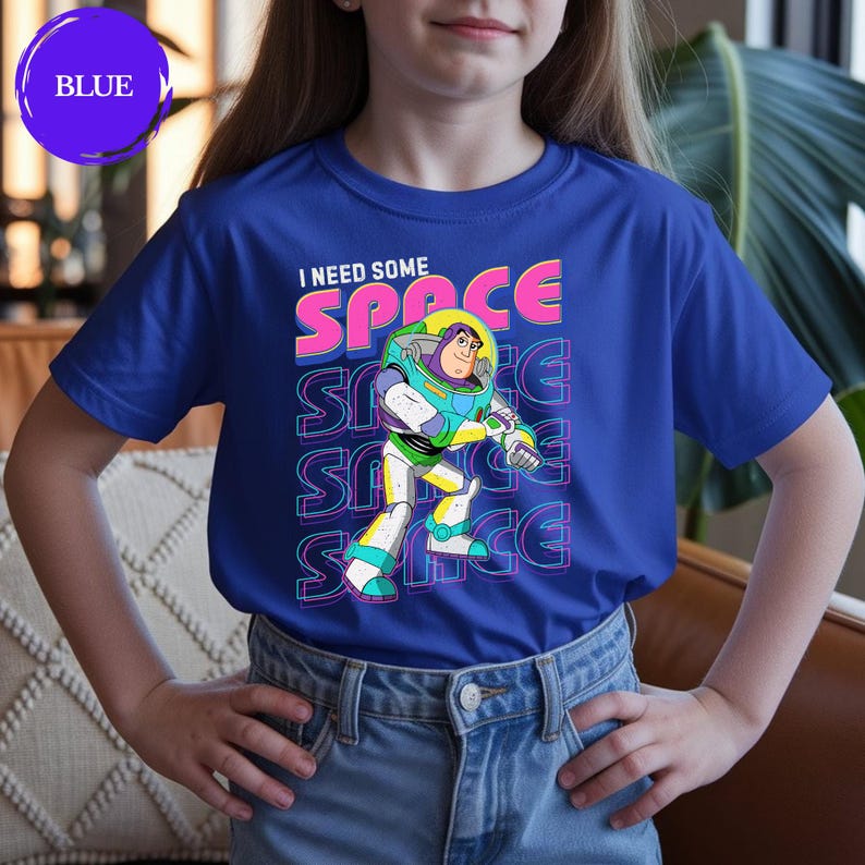 May include: Blue t-shirt featuring a graphic of Buzz Lightyear with the text "I NEED SOME SPACE". The design includes the word "SPACE" repeated vertically. The shirt is worn by a person.