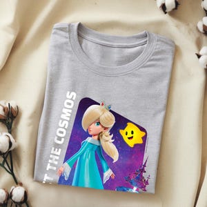 May include: A light gray t-shirt featuring a graphic of a character in a blue dress with a crown, a yellow star, and the text "'T THE COSMOS". The design is set against a purple and blue space background.