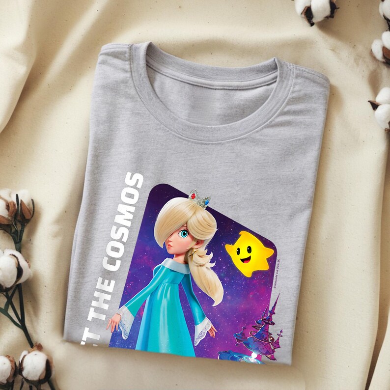 May include: A light gray t-shirt featuring a graphic of a character in a blue dress with a crown, a yellow star, and the text "'T THE COSMOS". The design is set against a purple and blue space background.