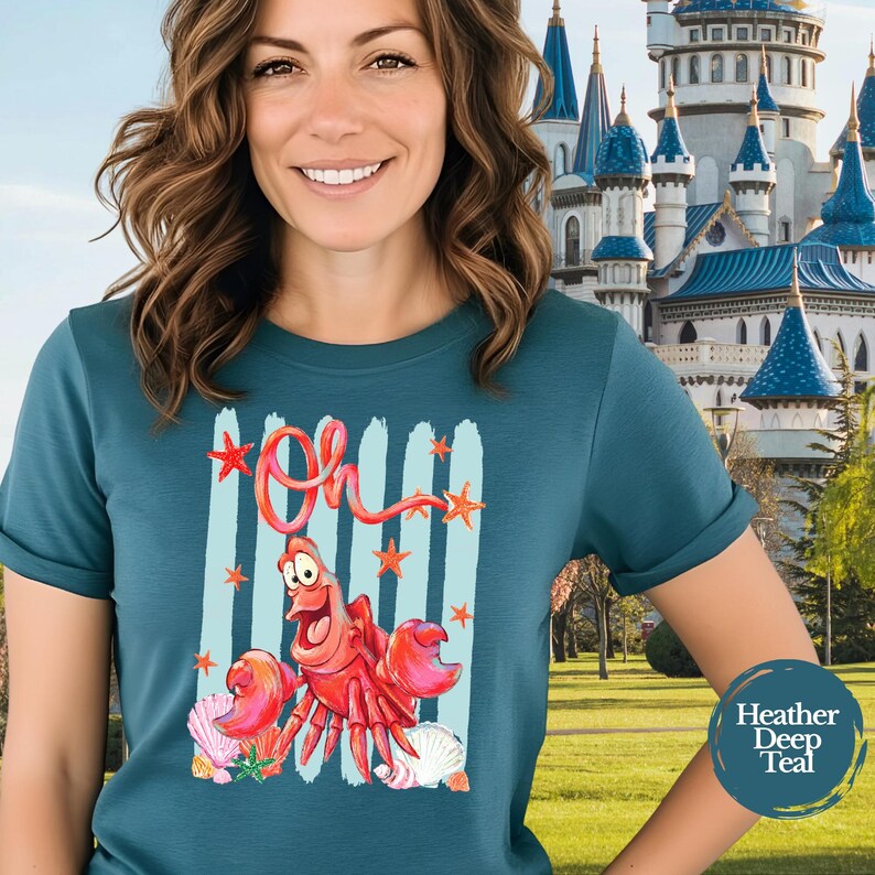 Pu&ograve; includere: Una t-shirt color verde acqua scuro m&eacute;lange con una stampa grafica di un granchio rosso e la parola Oh scritta in corsivo rosso sopra. Il design include pennellate verticali azzurre, piccole stelle marine rosse e varie conchiglie sul fondo.