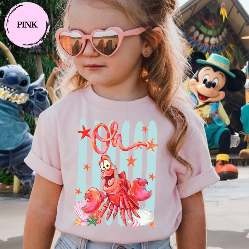 Pu&ograve; includere: Una t-shirt a maniche corte color rosa chiaro con una stampa grafica di un personaggio a forma di granchio rosso e il testo "Oh" in corsivo. Il design presenta pennellate verticali azzurre, stelle marine arancioni e conchiglie sul tessuto.