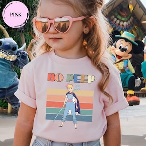 May include: A light pink t-shirt featuring a graphic of Bo Peep with a shepherd's crook, set against a background of horizontal stripes in muted colors. The text BO PEEP is printed in a bold, multi-colored font above the character illustration.