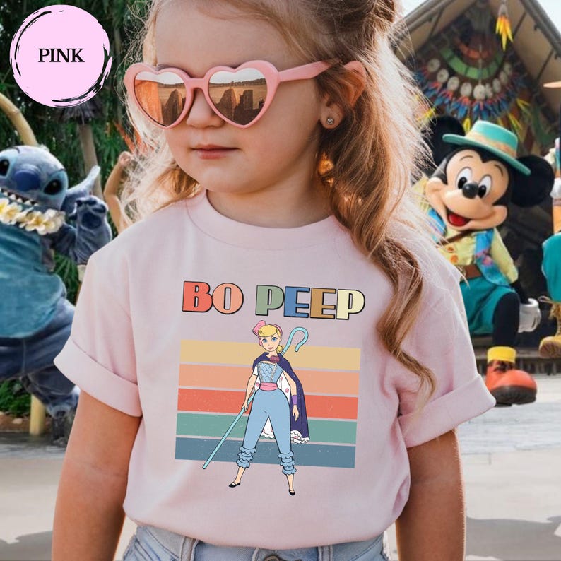 May include: A light pink t-shirt featuring a graphic of Bo Peep with a shepherd's crook, set against a background of horizontal stripes in muted colors. The text BO PEEP is printed in a bold, multi-colored font above the character illustration.