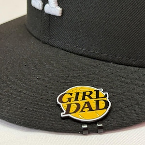 May include: Black baseball cap with a white embroidered "A" logo. A "GIRL DAD" pin with a basketball design is attached to the brim. The pin has a yellow background and black text.