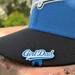 May include: A blue and black baseball cap with the text "Girl Dad" in white and blue lettering. The cap has a curved brim and a structured crown. The hat is resting on a brick surface.