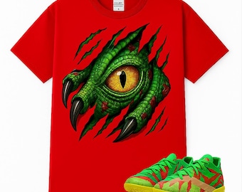 May include: A bright red short-sleeved t-shirt featuring a graphic of a green reptilian eye and clawed hand emerging from a torn fabric effect. Below the shirt are green and red athletic sneakers with yellow soles and a textured pattern.