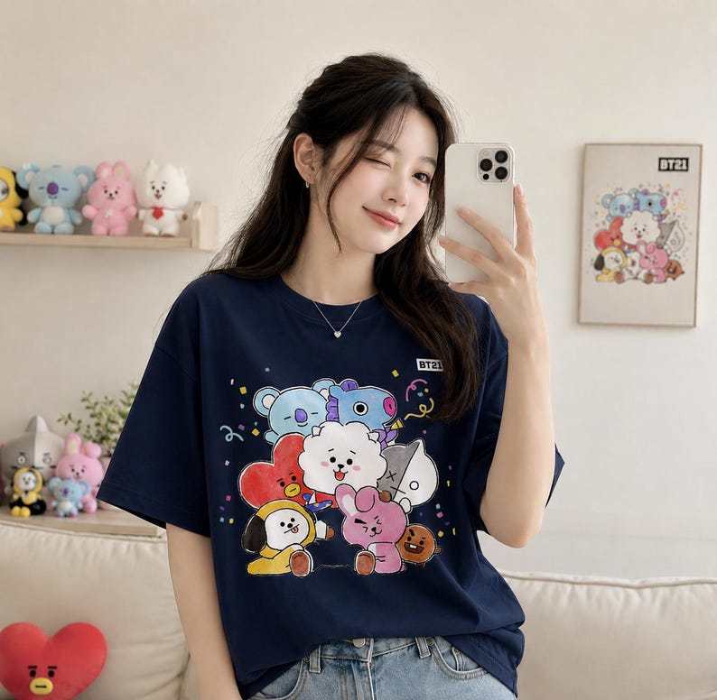 May include: Navy blue t-shirt featuring a colorful graphic of BT21 characters. The design includes cartoon figures in various colors, such as blue, pink, and yellow. The shirt has short sleeves and a crew neck.