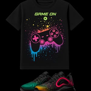 Pode incluir: Uma t-shirt preta de manga curta com o texto GAME ON e um gráfico colorido de um comando de videojogos com efeitos de salpicos de tinta. Por baixo da t-shirt estão sapatilhas pretas com um gradiente de tons verde, laranja e rosa.