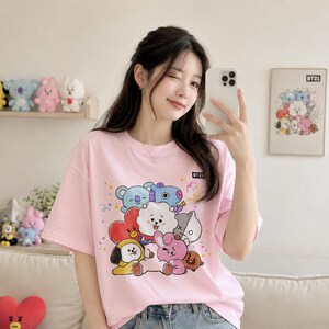 May include: A light pink t-shirt featuring a colorful graphic of cartoon characters. The shirt has short sleeves and a crew neck. The text "BT21" is printed on the shirt. The shirt is worn by a person.