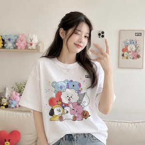 May include: White t-shirt featuring a colorful graphic of cartoon characters. The shirt has short sleeves and a crew neck. The graphic includes various characters in different colors. The shirt is worn by a person.