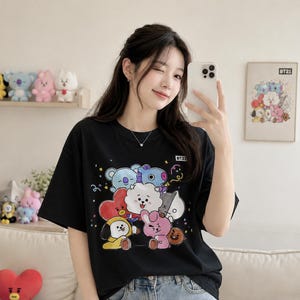May include: Black t-shirt featuring a colorful graphic of cartoon characters. The shirt has a crew neck and short sleeves. The graphic includes various characters in shades of blue, pink, yellow, and white. The shirt has the text "BT21" on the upper right.