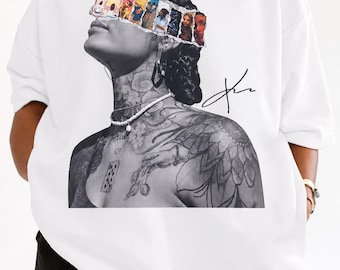 May include: White sweatshirt featuring a black and white portrait with colorful images across the eyes. The portrait has a signature in black ink. The model is wearing a gold necklace and a pearl necklace.