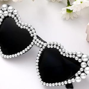 May include: Heart-shaped sunglasses with black lenses and a frame embellished with faux pearls. The sunglasses have a classic design with a modern twist, perfect for adding a touch of glamour to any outfit.