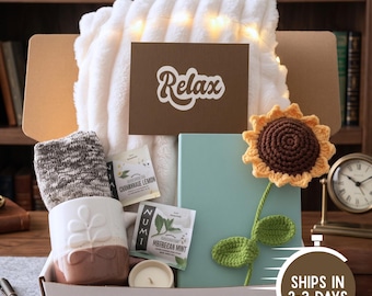 May include: A gift box containing a white faux fur throw, a brown card with the word Relax, a pair of grey knit socks, a white ceramic mug with a leaf pattern, two Numi tea bags, a small candle, a light blue notebook, and a crocheted sunflower.