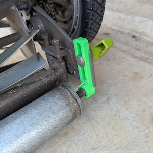 May include: Close-up of a lawn mower mechanism. The image shows a silver metal roller, blades, and a wheel. A bright green plastic component is attached to the mower, with a smaller yellow-green piece nearby. The mower appears to be used.