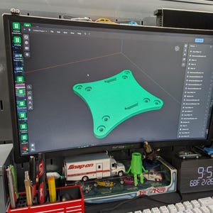 Könnte beinhalten: Ein Computermonitor zeigt ein 3D-Modell eines grünen, vierseitigen Objekts mit abgerundeten Ecken und Löchern. Der Bildschirm zeigt eine Designprogrammoberfläche. Eine Digitaluhr zeigt Zeit, Datum und Temperatur an. Spielzeugautos und ein Snap-on-Truck-Modell stehen auf dem Schreibtisch.
