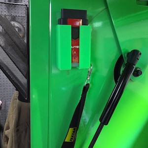 Puede incluir: Un soporte de plástico verde brillante con un dispositivo negro y rojo en su interior, montado sobre una superficie verde. Una correa negra con texto blanco cuelga debajo. El dispositivo tiene la palabra "UTE" visible.