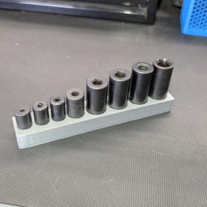 May include: A set of eight black metal sockets of varying sizes, arranged in a row on a light gray holder. The sockets are cylindrical with a threaded interior. The holder is rectangular and appears to be 3D-printed.