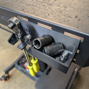 Può includere: Un organizer per utensili nero stampato in 3D attaccato a un banco da lavoro. L'organizer contiene vari utensili, tra cui bussole, cricchetti e altro hardware. Il banco da lavoro è grigio scuro e gli utensili sono argentati e neri.