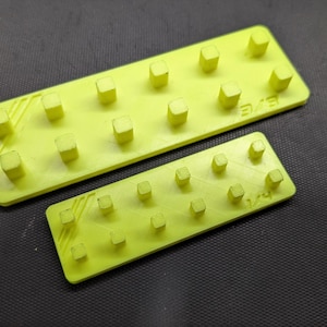 May include: Two neon yellow rectangular plastic pieces with raised square pegs. The larger piece has 12 pegs, and the smaller has 6. The pieces are on a dark gray textured surface. The larger piece has the text "EVE" on it.