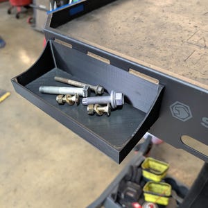 Può includere: Un tavolo da lavoro in metallo nero con un vassoio integrato contenente vari bulloni e dadi. Il vassoio è angolato e posizionato sul lato del tavolo. Il tavolo presenta un logo sul lato.