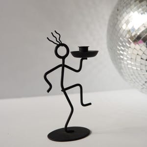 May include: A black metal candle holder shaped like a stick figure in a dancing pose. The figure has a circular head with three wavy lines on top, bent arms and legs, and holds a small circular tray for a candle. It stands on a flat oval base.