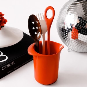 May include: A bright orange cylindrical kitchen utensil holder with a pouring spout rim. Inside the holder are three utensils: a dark brown slotted spoon, a white fork, and a long-handled orange spoon with a circular opening at the top.