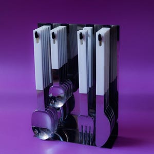 Può includere: Un set di posate in acciaio inossidabile con manici bianchi e dettagli neri, esposto in un supporto cromato. Il set include cucchiai, forchette e coltelli, su uno sfondo viola. Gli utensili sono riflettenti, creando un'estetica moderna.