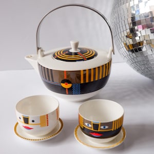 May include: A ceramic teapot with a metal handle and two matching teacups on saucers. The set features a cream base with black, brown, yellow, and red geometric patterns, including stylized facial features on the cups and circular motifs on the lid.