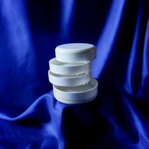 May include: A white plastic storage organizer featuring four circular, tiered compartments that rotate around a central axis. The structure consists of a solid top lid and three open, shallow trays stacked vertically in a cylindrical shape.