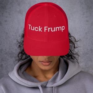 May include: A red trucker hat with white embroidered text that reads "Tuck Frump". The hat has a mesh back and a curved brim. The person wearing the hat is wearing a gray hoodie.