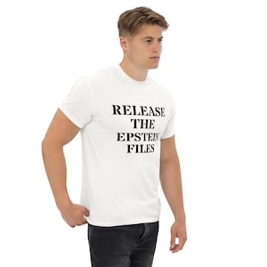 Puede incluir: Camiseta blanca con el texto "RELEASE THE EPSTEIN FILES" en negro y negrita. La camiseta es de manga corta y parece estar hecha de un material suave. La persona que lleva la camiseta está de pie y mirando hacia un lado.
