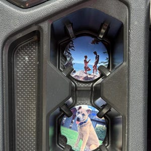 May include: Black car console with two custom car coasters. One coaster features a couple on a rocky beach, the other a tan dog with a blue collar. The coasters are round and fit into the cup holders.