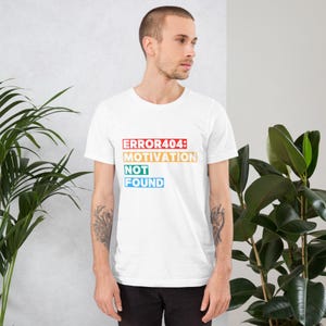 Può includere: T-shirt bianco con la scritta "ERROR404: MOTIVATION NOT FOUND" in un design multicolore a strati. La maglietta a maniche corte sembra essere realizzata in un materiale morbido. Il testo è in rosso, giallo, verde e blu.