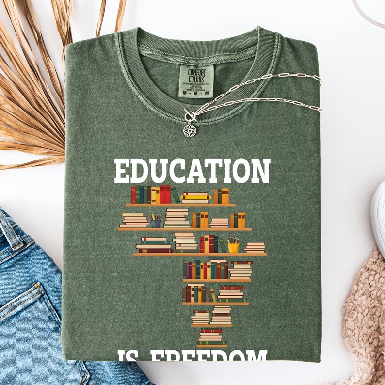 May include: Olive green t-shirt with the word "EDUCATION" in white block letters. A graphic of stacked books on shelves forms a stylized shape. The text "IS FREEDOM" is partially visible at the bottom. A silver necklace is draped across the shirt.