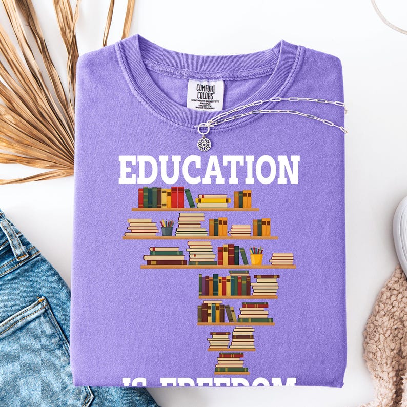May include: A lilac Comfort Colors sweatshirt with the word "EDUCATION" in white block letters. Below the word is a graphic of bookshelves filled with books. A silver necklace with a sunflower pendant is draped across the shirt.