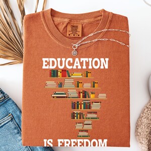 May include: A rust-colored t-shirt with the words "EDUCATION IS FREEDOM" in white. The shirt features a graphic of bookshelves filled with colorful books. A silver necklace with a pendant is draped across the shirt.