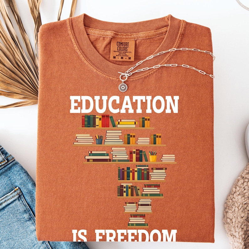 May include: A rust-colored t-shirt with the words "EDUCATION IS FREEDOM" in white. The shirt features a graphic of bookshelves filled with colorful books. A silver necklace with a pendant is draped across the shirt.