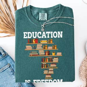 May include: A teal Comfort Colors sweatshirt with the word "EDUCATION" in white block letters. Below the word is a graphic of stacked bookshelves with colorful books. The phrase "IS FREEDOM" is at the bottom. A silver necklace is draped across the shirt.