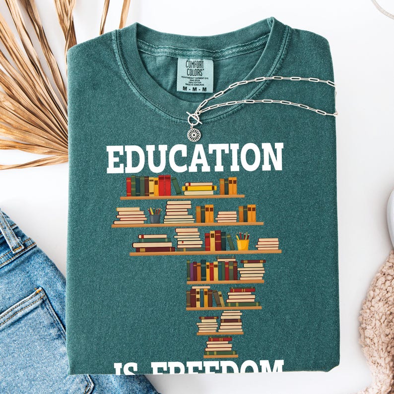 May include: A teal Comfort Colors sweatshirt with the word "EDUCATION" in white block letters. Below the word is a graphic of stacked bookshelves with colorful books. The phrase "IS FREEDOM" is at the bottom. A silver necklace is draped across the shirt.