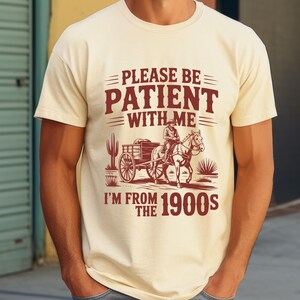 May include: Cream-colored t-shirt with a vintage-style graphic. The graphic features a man on horseback pulling a cart, with text that reads "PLEASE BE PATIENT WITH ME I'M FROM THE 1900s" in a rustic font.