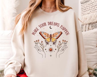 May include: Cream-colored sweatshirt with the text "MAKE YOUR DREAMS HAPPEN" and a butterfly graphic. The butterfly is orange and yellow with brown accents. The design includes hands, flowers, a sun, and stars.
