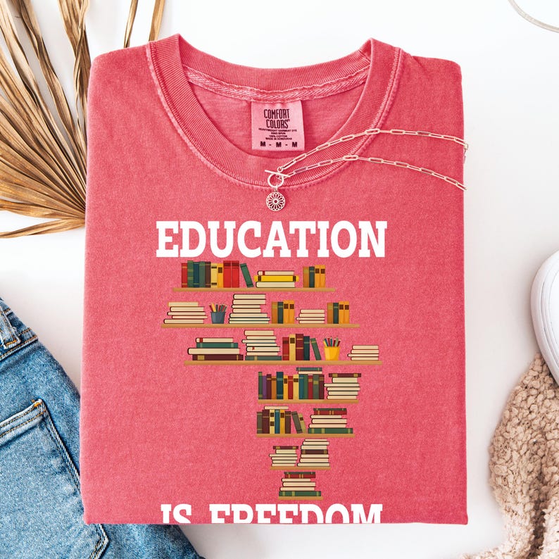 May include: A red t-shirt with the words "EDUCATION IS FREEDOM" in white. The design features a graphic of stacked books on shelves. A silver necklace with a pendant is draped over the shirt.
